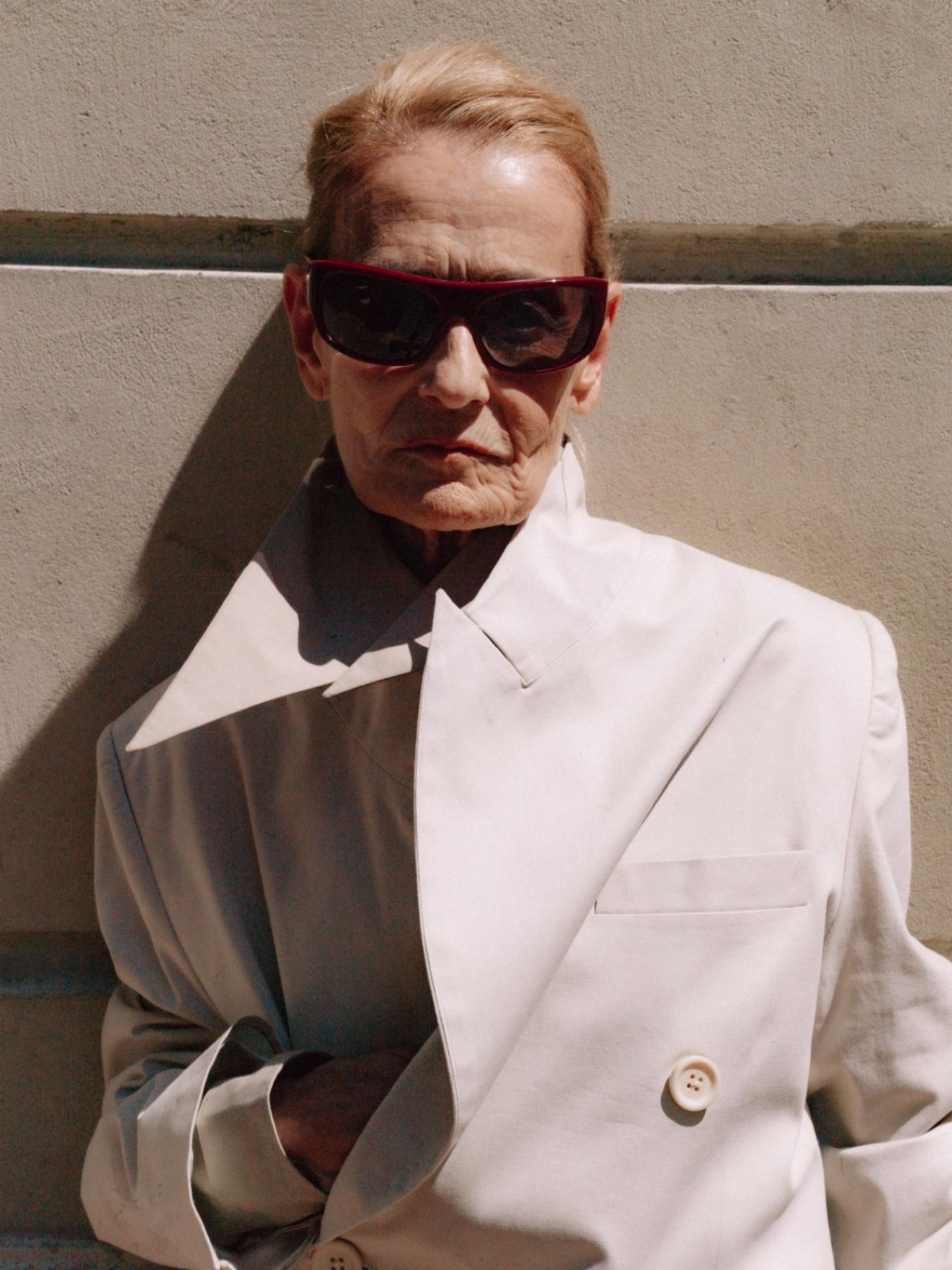 A woman in dark sunglasses and a white coat against a stone wall