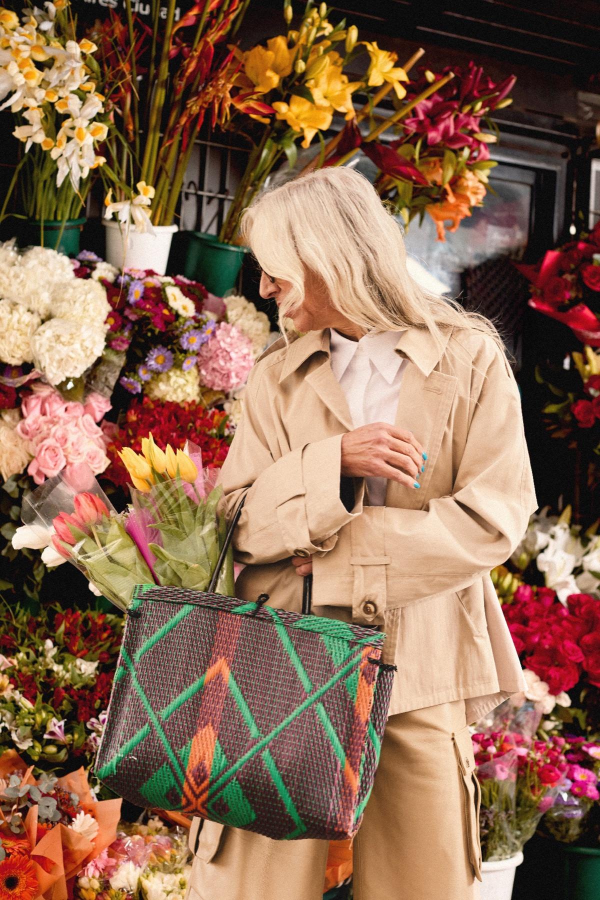 A woman in a trench coat choosing flowers at a street stall, a woven bag on her arm