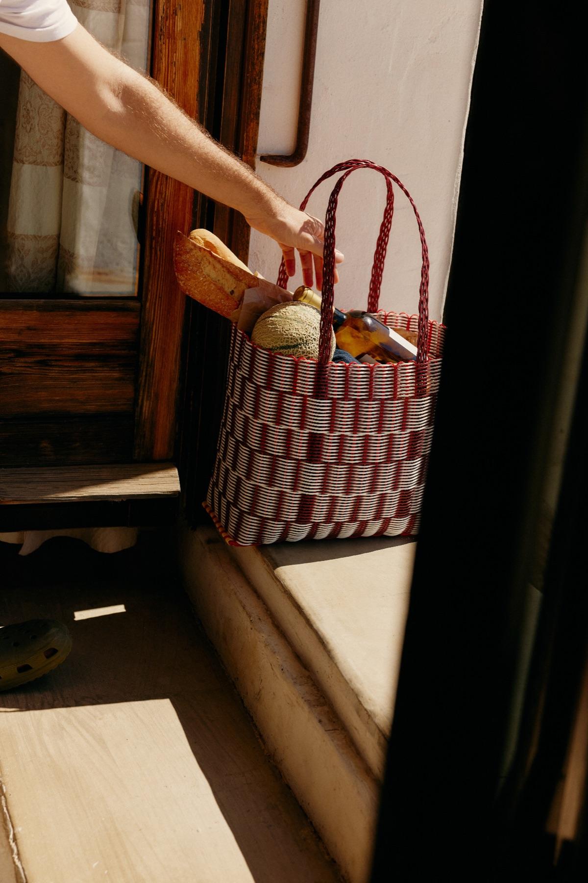 A hand setting a woven shopping bag full of groceries on a sunny windowsill