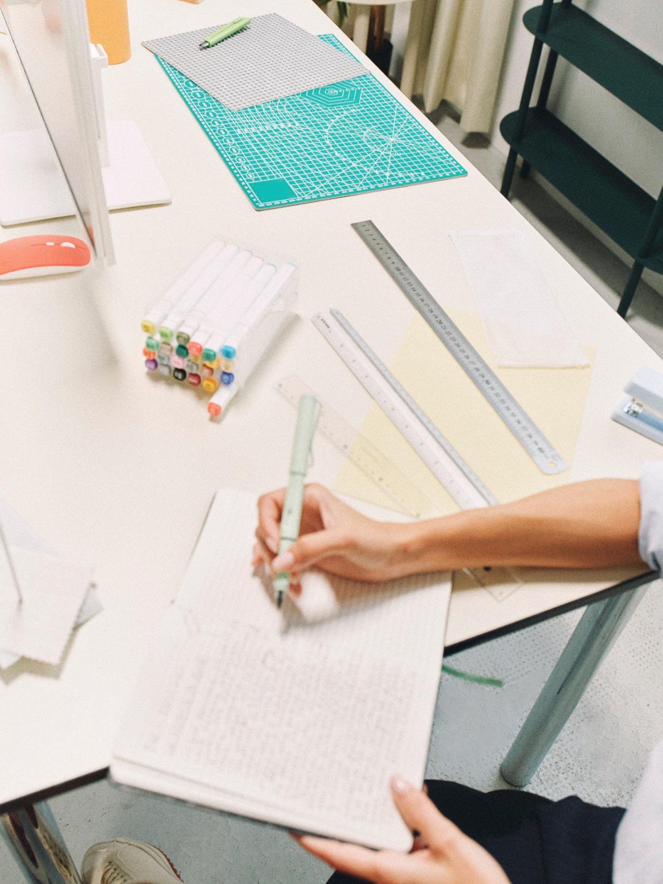 A hand sketching page layouts in a notebook beside a ruler and cutting mat
