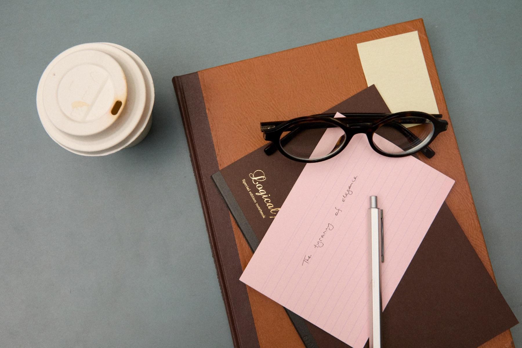 A notebook, glasses, a pen and a coffee cup on a gray desk