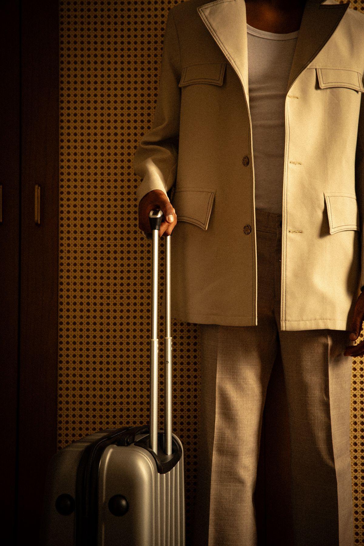 A traveler in a linen suit resting a hand on a suitcase