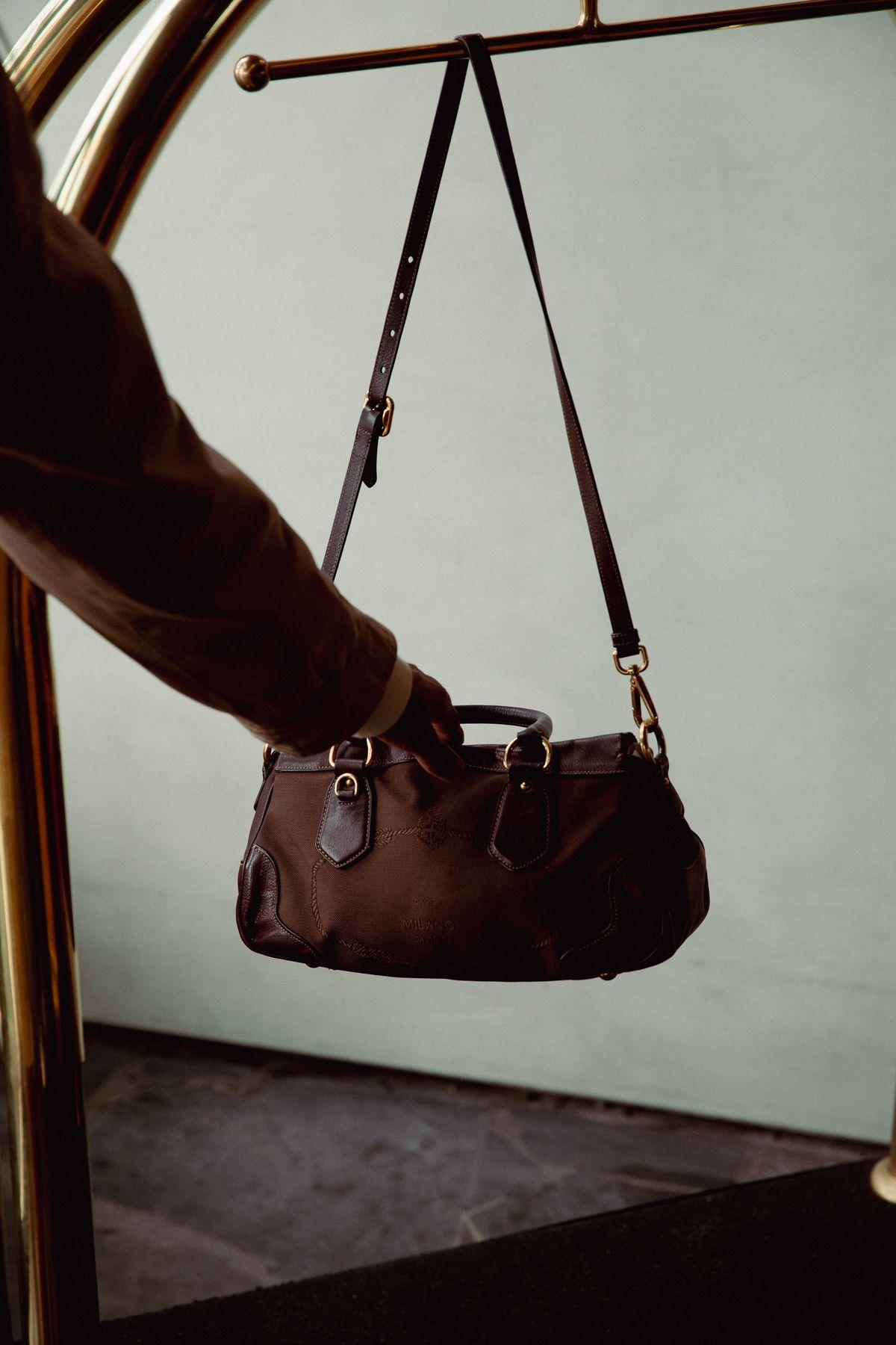 A hand hanging a leather bag on a brass luggage cart